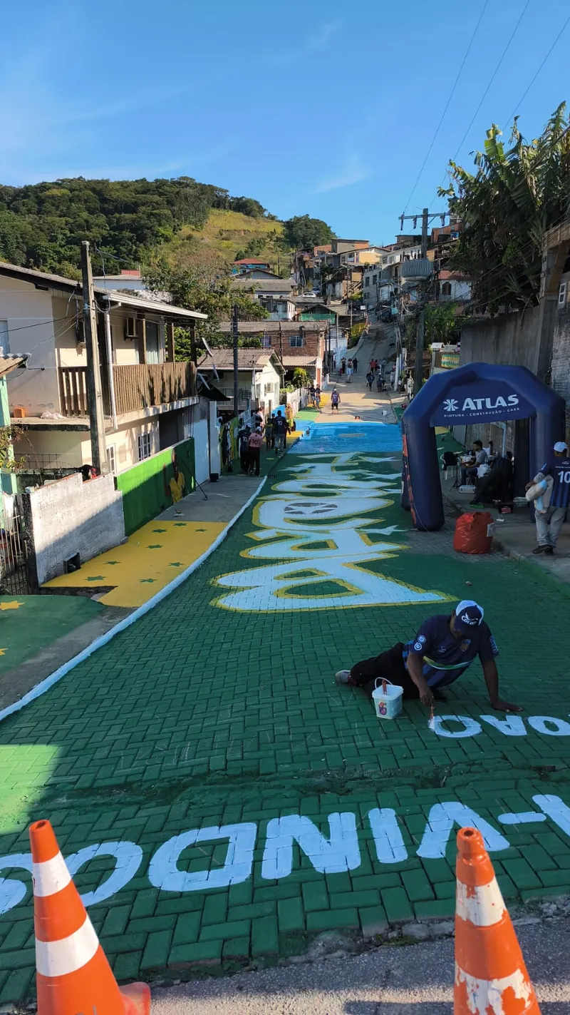 Rua de comunidade com o calçamento pintado de verde e amarelo durante o mutirão Rumo à Copa, com pintores trabalhando e tenda da Atlas ao fundo